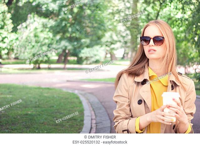 Young fashionable woman taking a coffee break after shopping, walking with a coffee-to-go in her hands against green city park background