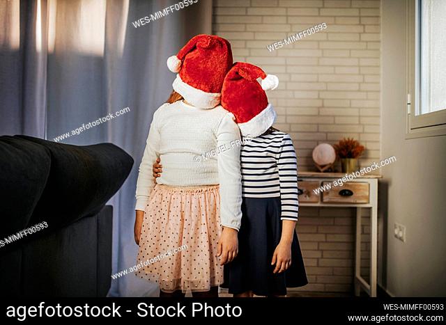 Sisters covering their faces with Santa hat while standing in living room