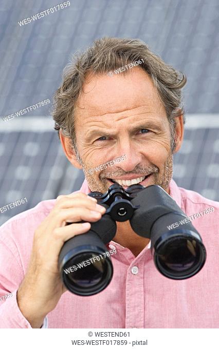 Germany, Munich, Mature man holding binocular in solar plant, smiling, portrait