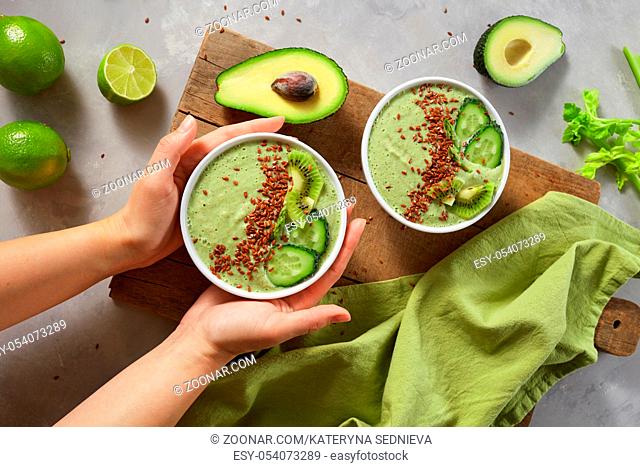 Fresh cucumber avocado smoothie. Female hands holding a plate of vegetable smoothies with kiwi and flax seeds against a wooden board. Top view
