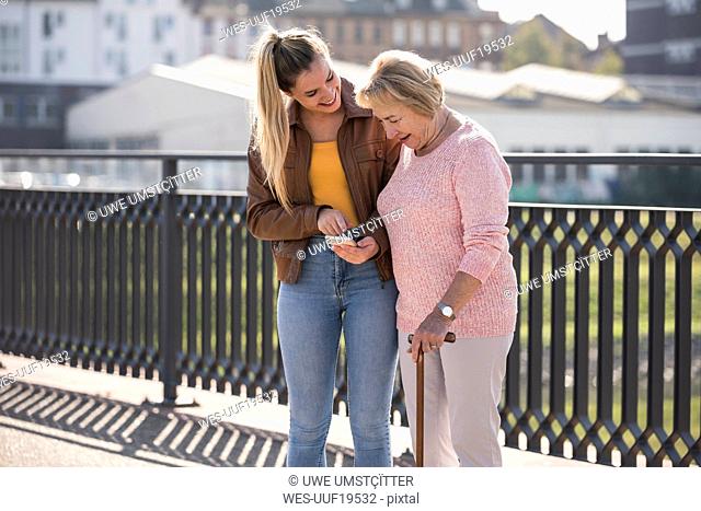 Granddaughter and her grandmother walking on footbridge and using smartphone
