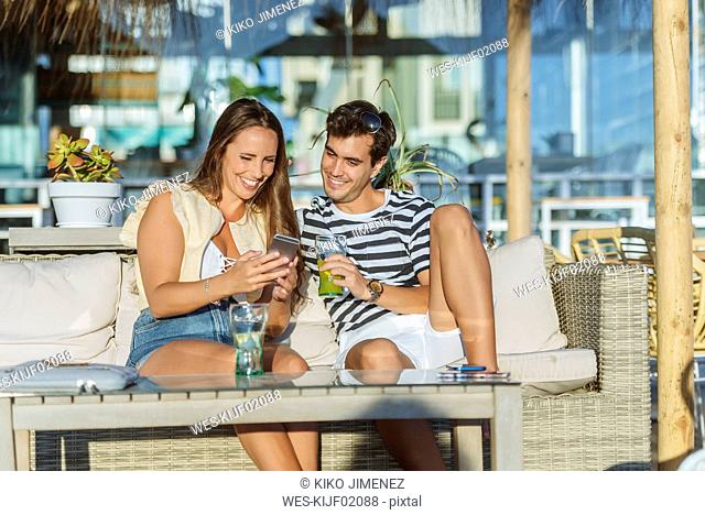 Happy young couple sitting on the terrace of a bar looking at cell phone