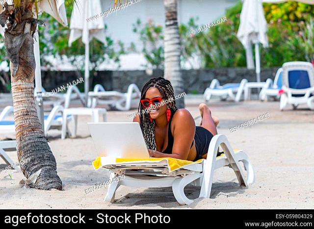 African-American woman on the beach relaxing in deck chair using laptop looking at camera. Cheerful adult woman enjoying a summer day at the beach