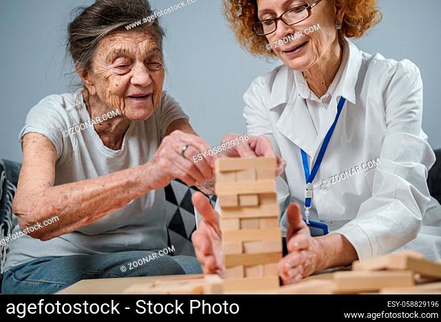 Dementia therapy in playful way, training fingers and fine motor skills, build wooden blocks into tower, playing Jenga. Senior woman 90 years old and doctor...