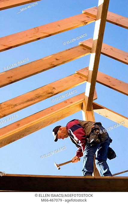 Carpenter hammering trimmer to center post while standing below the framing of the roof rafters at a construction site