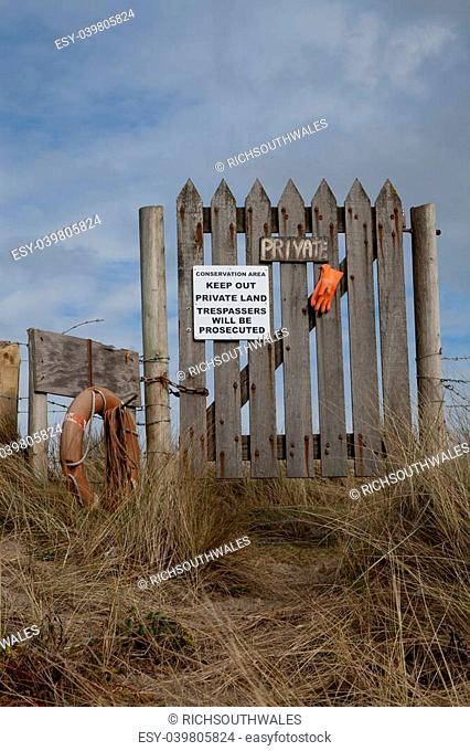 A wooden gate and fence with a 'private' sign and warning 'trespassers will be prosecuted' on a sand dune against a blue sky with cloud