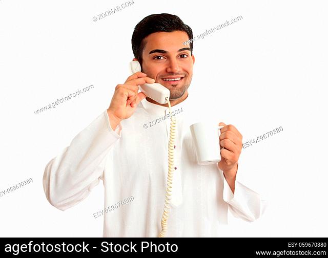 A friendly smiling ethnic arab businessman on a telephone call. He is holding a mug of coffee and is wearing traditional middle eastern or south east asian...