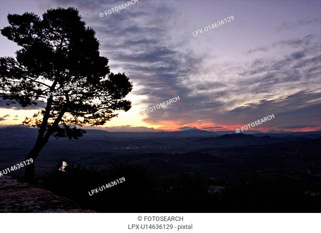 Evergreen tree silhouetted beside old stone town wall at sunrise, with view over Tiber valley in misty dawn of winter, river winding through valley