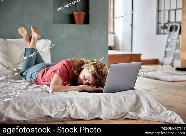 Tired woman lying by laptop on bed at home