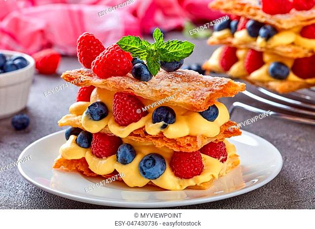 french dessert millefeuille of three pieces of puff pastry layered with custard cream, raspberries, blueberries on a white plate on a concrete table
