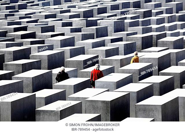 Three people dressed in red, black and yellow-gold jackets walk amongst concrete slabs of the Holocaust Memorial, Berlin, Germany