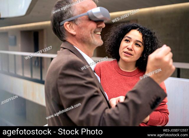 Businesswoman looking at happy colleague wearing VR glasses in corridor