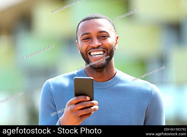 Front view portrait of a happy man with black skin laughing holding phone looking at you in the street