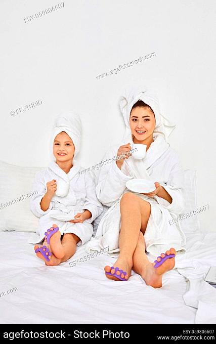 happy woman and her daughter in white bathrobes and towels on heads with pedicure sponge on fingers drinking tea