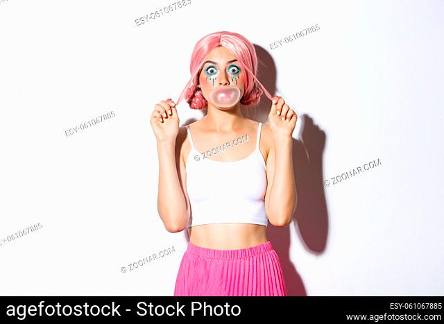 Portrait of attractive girl blowing pink bubble gum on face and looking silly, standing in anime wig and party outfit