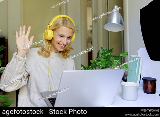 Smiling woman wearing headphones waving on video call over laptop at home