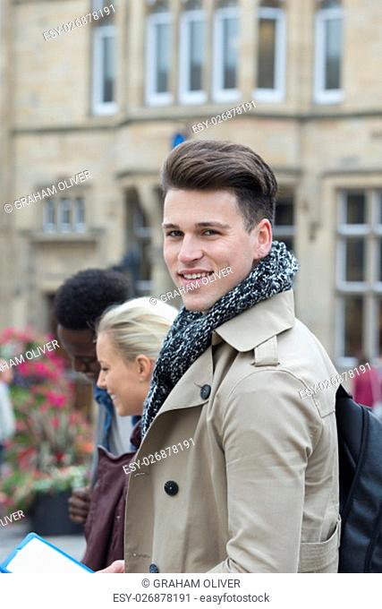 A young male looks at the camera smiling as he stands outside in the city centre with friends. He is wrapped up in a coat and scarf for the cold weather