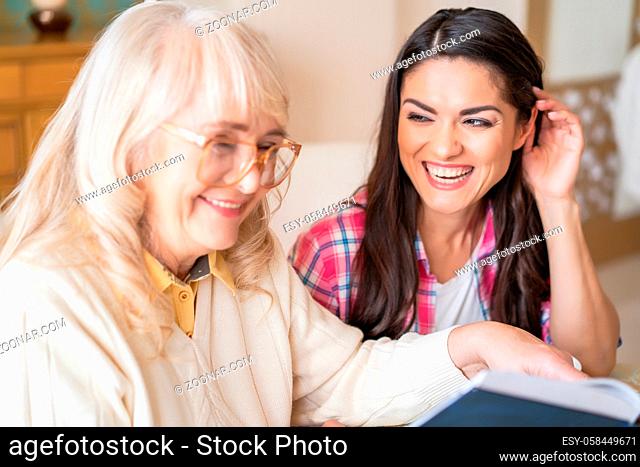 Two Wonderful Women Laugh Over Funny Story They Have Read From A Book. Daughter Looks With Love And Care On Her Senior Mother In Glasses Smiling