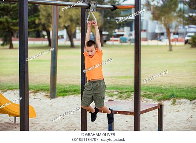 A side-view shot of a young caucasian boy wearing casual clothing, he is swinging from a metal frame in a public park