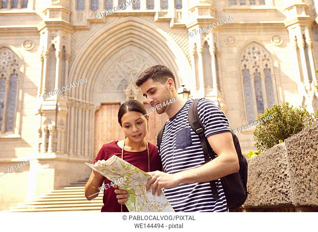 Young tourist couple on vacation with a backpack, looking at a map while dressed casually in jeans and t-shirts with a cathedral behind them