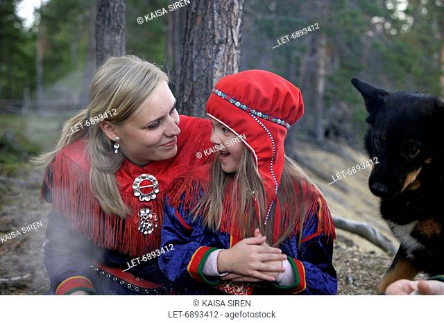 A Sami mother and a daughter dressed in the traditional Lapp dress and a Lapland reindeer dog are having a picnic by open fire in Inari