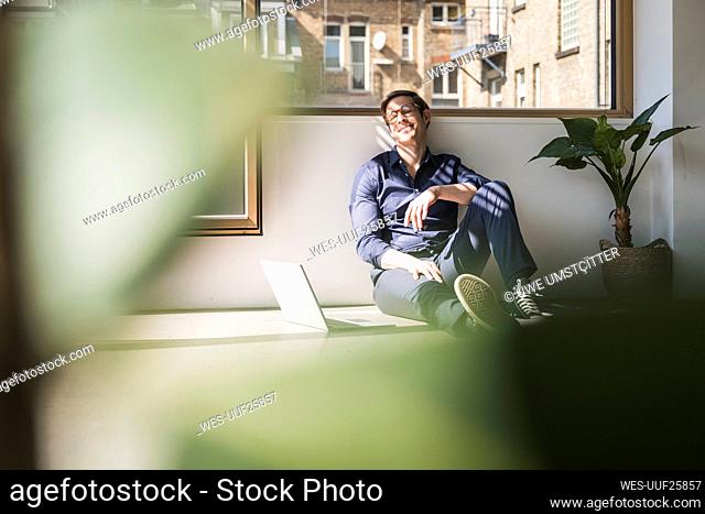 Relaxed businessman sitting on the floor in office next to laptop