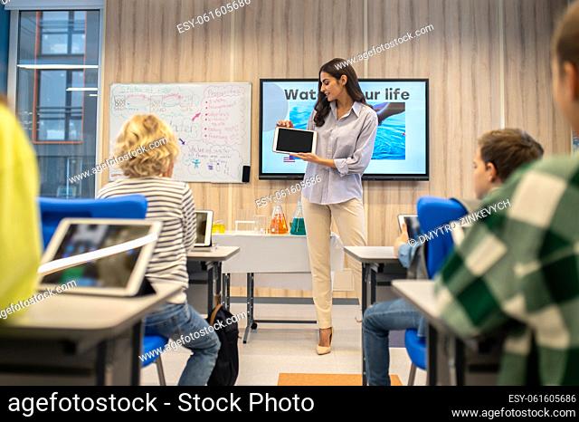 Smart school. Young pretty woman showing tablet screen to children sitting at lesson at desks in chemistry class