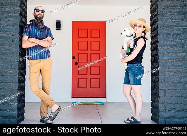 Smiling woman carrying dog while man with arms crossed standing in front of house