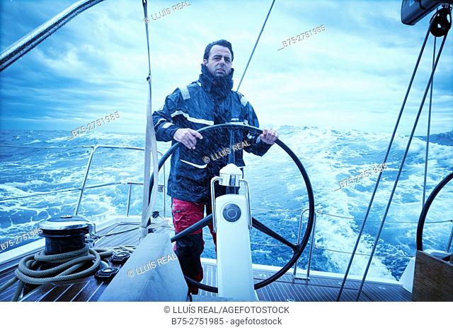 Man dressed in waterproof clothing at the helm of sailboat riding, rough sea. Mediterranean sea