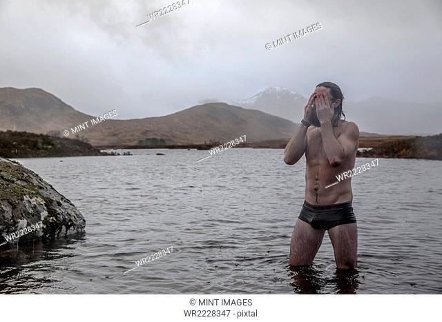 A man standing thigh deep in sea water in swimming trunks wiping water from his eyes