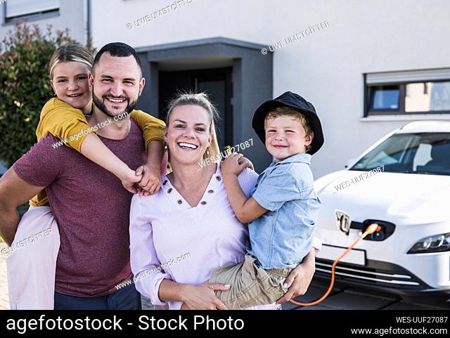 Happy family standing in front of family home and electric car