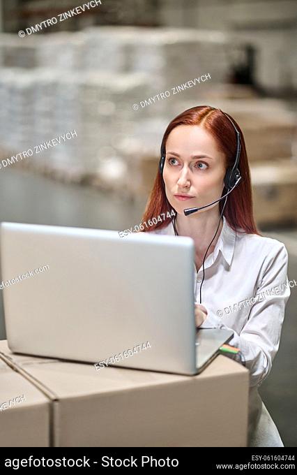 Working process. Focused young woman in headset working on laptop looking up and away standing in warehouse