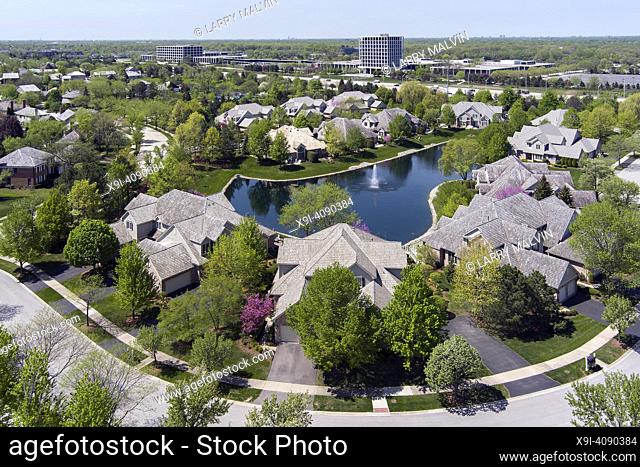 Aerial view of a neighborhood community in a suburban setting with a pond and highway with an office highrise in the distance.