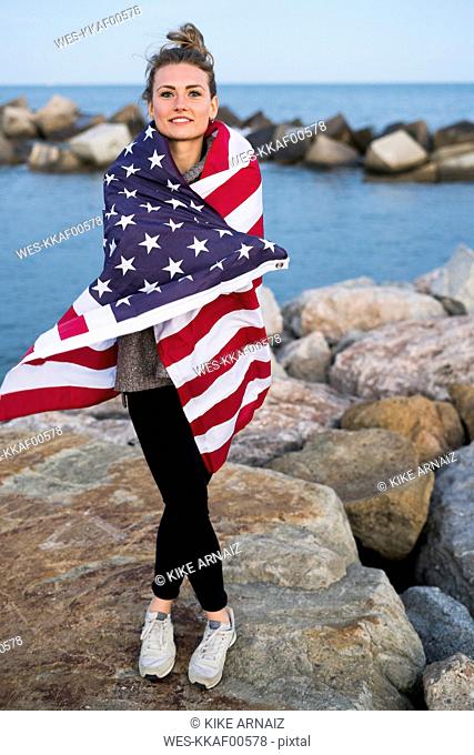 Portrait of young woman standing on rocks in front of the sea wrapped in US American flag