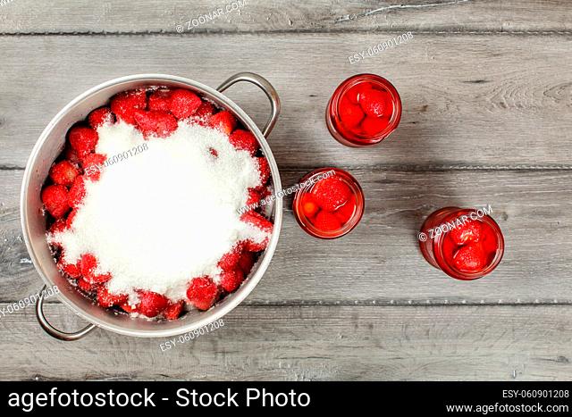 Tabletop view, large steel pot full of strawberries covered with crystal sugar, three bottles of strawberry in syrup next to it on gray wood desk