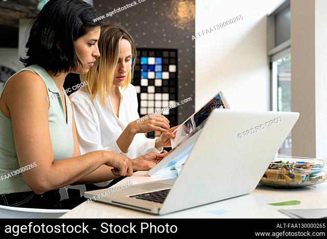 Side view shot of two female interior designers at their desk working with books and a laptop computer