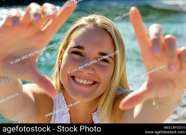 Cheerful young woman showing finger frame while standing against lakeshore on sunny day