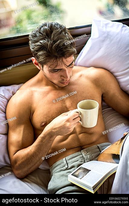 Sexy handsome young man laying shirtless on his bed next to window, holding a coffee or tea cup while reading a book