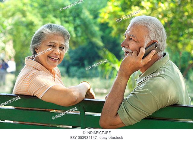 Senior man talking on mobile phone at park with woman smiling at camera
