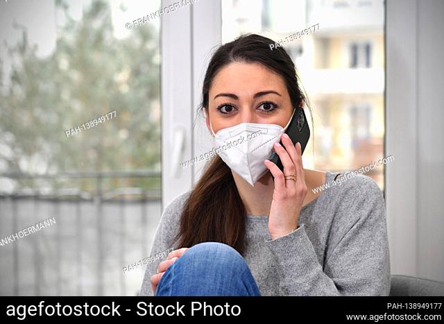 A young woman is sitting in her apartment, working from home because of the corona pandemic, wearing an FFP2 mask and talking on the phone with a smartphone