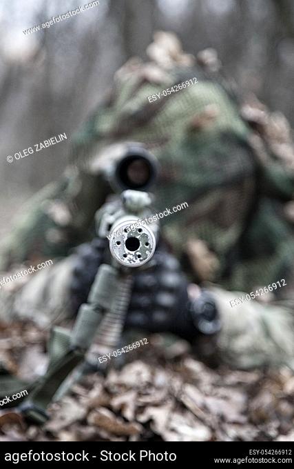 Close up image of sniper rifle silencer muzzle, front view. Army elite forces shooter lying on ground in forest. Commando sniper hiding