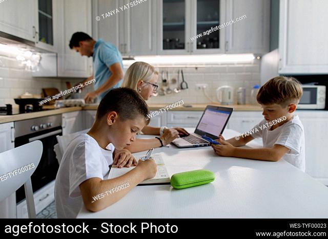 Boy studying on table with family in background at home