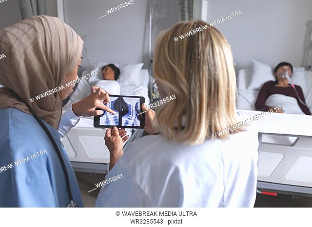 Rear view of diverse female doctors discussing over x-ray report on digital tablet in the ward at hospital. Diverse female patients are sleeping in bed in the...