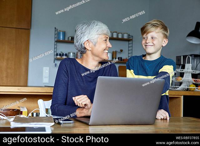 Smiling grandson standing next to grandmother working on laptop at home