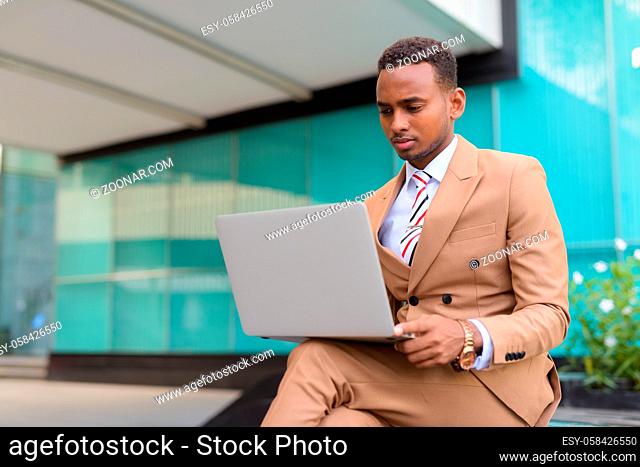 Portrait of young handsome African businessman wearing suit outside modern building in the city