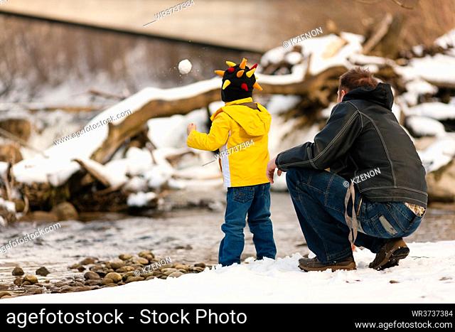 Family - father and son to be seen - on a walk along a riverbank in winter; the child is throwing a snowball