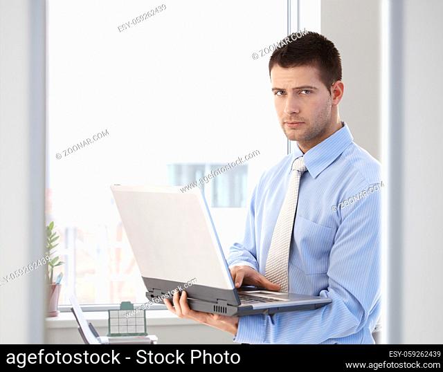Young handsome businessman working on laptop, standing in bright office
