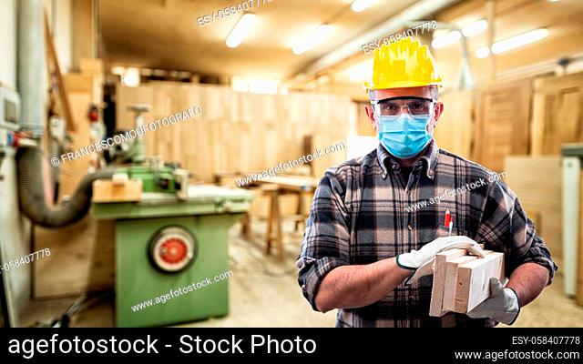 Carpenter worker at work in the carpentry workshop, wears helmet, goggles, leather gloves and surgical mask to prevent coronavirus infection