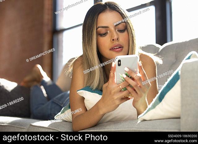 young Hispanic woman text messaging through mobile phone while lying on sofa in living room at home