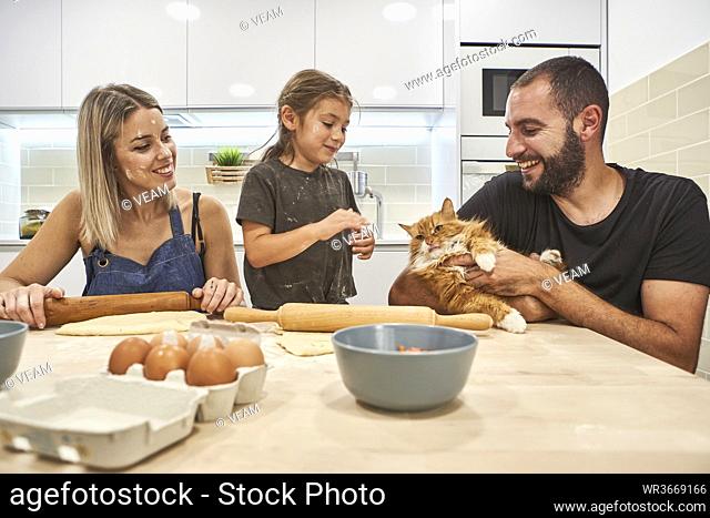 Mother kneading dough with rolling pin while father and daughter playing with cat on kitchen's table